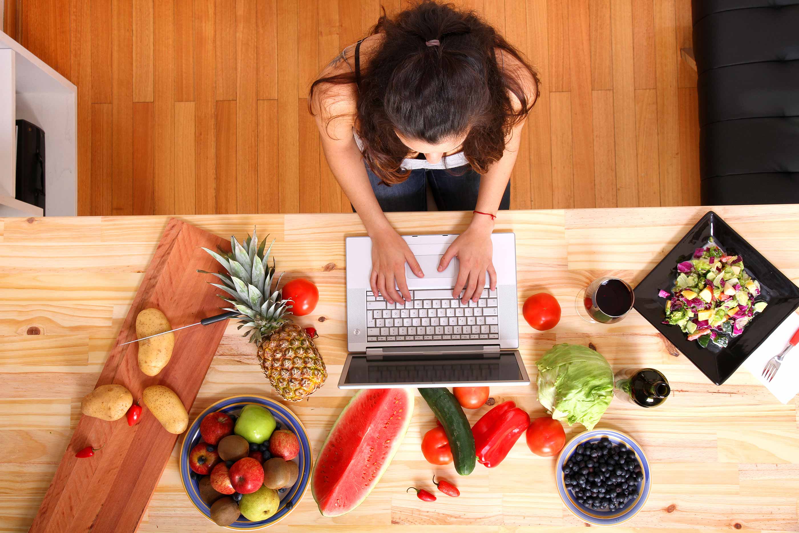 Woman researching recipes on a laptop, on a table surrounded by fresh produce.